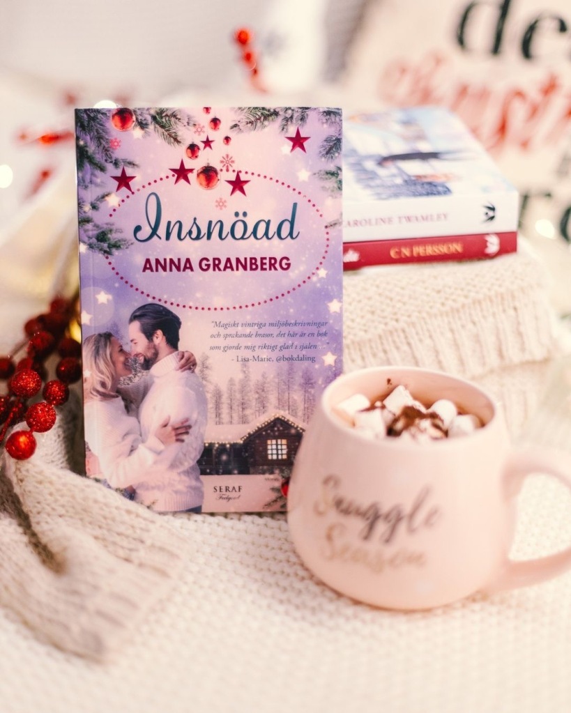 Boken Insnöad står uppställd mot två andra nyutkomna julromaner. I förgrunden syns en kopp varm choklad med marshmallows.